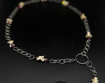 May include: A necklace featuring a silver-toned metal chain interspersed with small, off-white bone segments and dark purple faceted beads. The chain connects to a large circular ring, from which hangs a black heart-shaped pendant with a white graphic.