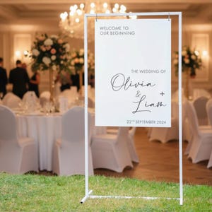 Könnte beinhalten: Ein weißes Hochzeitszeichen aus Metall. Der Text lautet "WELCOME TO OUR BEGINNING" oben, gefolgt von "THE WEDDING OF Olivia & Liam" und dem Datum "22nd SEPTEMBER 2024". Das Schild steht auf Gras, in einem Hochzeitsort.
