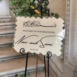 Op de afbeelding: Een helder acryl trouwbord met zwarte kalligrafie. Het bord leest "Welcome to Our Beginning" en bevat de namen "Noor and Sami" met de datum 6/6/25. Het bord wordt weergegeven op een zwart metalen ezel.