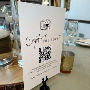以下が含まれることがあります： 「Capture the Love」の文字とQRコードが印刷された白いカード。黒いスタンドに立てられています。レンズにハートのマークが入ったカメラのイラストが特徴です。写真の共有を促すテキストも含まれています。