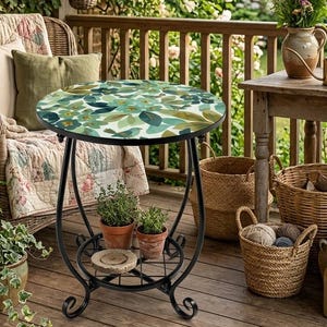 May include: A small, round side table with a black wrought iron frame and a decorative top. The tabletop features a leaf pattern in shades of green and blue. The table has a lower shelf for storage and is surrounded by plants and outdoor decor.