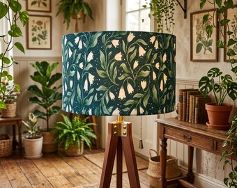 May include: A dark green lampshade with a white floral pattern sits atop a wooden tripod lamp base. The lamp is in a room with wooden floors, plants, and botanical prints, creating a cozy, natural aesthetic.