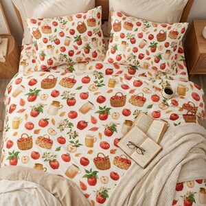 Pode incluir: Um conjunto de roupa de cama com fundo creme e um padrão repetitivo de maçãs vermelhas, cestos de vime cheios de maçãs, flores de macieira brancas e jarros de cerâmica bege. Um livro aberto e óculos repousam sobre a colcha.