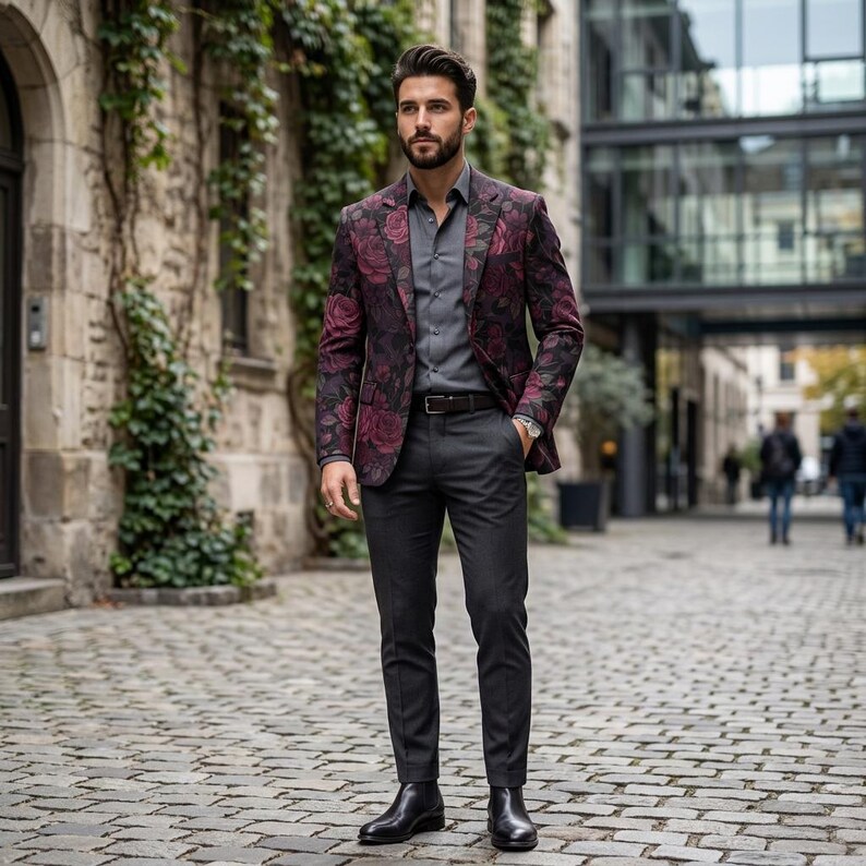 May include: A man's suit featuring a burgundy blazer with a floral pattern, a gray dress shirt, and dark gray trousers. The suit is paired with a black belt and black boots. The background is a cobblestone street.