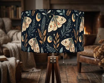 May include: A cylindrical lamp shade featuring a dark navy blue background with a repeating pattern of cream-colored moths, golden crescent moons, small stars, and various green and brown botanical leaf sprigs, supported by a wooden tripod base.