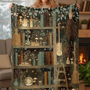 May include: A decorative throw blanket with a whimsical bookshelf design. The blanket features a dark background with illustrations of books, plants, mushrooms, and a ladder. The colour palette includes shades of teal, brown, and gold.