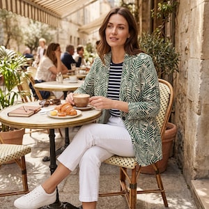 May include: A woman in a green leaf-print blazer and striped top, seated at a cafe table with a coffee cup and croissant. She wears white trousers. The scene is set outdoors with other people and a stone wall in the background.