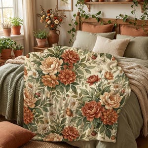 May include: A soft throw blanket featuring a repeating botanical pattern of large orange, peach, and cream-colored dahlias and small white daisies with green foliage on a light cream background, draped over a bed with pillows and bedding.