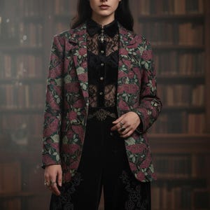 May include: A dark blazer featuring a floral design of burgundy roses and green leaves. The blazer is worn over a black lace top and black velvet trousers. The outfit is accessorized with rings.