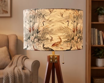 May include: A floor lamp with a cylindrical fabric shade featuring a printed pattern of cranes, koi fish, bamboo, and flowers in muted tones. The lamp has a dark wood tripod base with brass-colored metal hardware and a gold-toned pull chain switch.