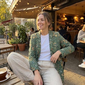 May include: A woman wearing a green floral blazer over a white t-shirt and tan pants, seated at an outdoor cafe. The blazer features a detailed botanical pattern in shades of green, tan, and cream. The cafe setting includes wooden tables, potted plants, and string lights.