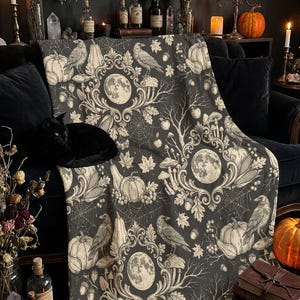Peut inclure: Un plaid gris foncé avec un motif répétitif d'illustrations blanches comprenant des pleines lunes, des corbeaux, des citrouilles, des champignons, des glands, des feuilles d'érable et des toiles d'araignée, entourés de volutes ornementales.