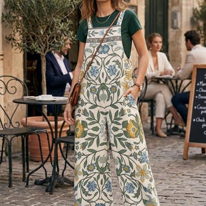 Könnte beinhalten: Eine Frau trägt einen cremefarbenen Jumpsuit mit einem Blumenmuster in Blau, Gelb und Grün. Der Jumpsuit hat weite Beine und Träger. Darunter wird ein grünes T-Shirt getragen, und sie trägt eine braune Tasche. Im Hintergrund eine europäische Straßenszene.