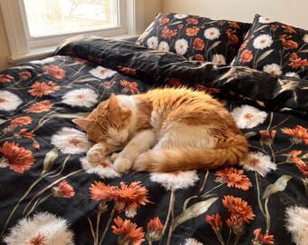 May include: A ginger and white cat sleeping on a black duvet cover and matching pillowcases featuring a repeating pattern of orange flowers and white dandelion seed heads. The bedding set displays a dark background with botanical floral motifs.
