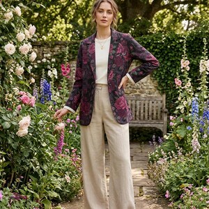 May include: A floral print blazer with a dark background and burgundy roses, paired with beige wide-leg pants and a white top. The outfit is completed with brown leather shoes. The setting is a garden with flowers and a stone path.