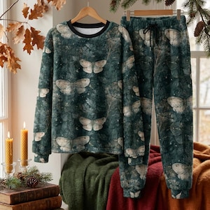 Op de afbeelding: Een donkergroene, pluizige set bestaande uit een sweatshirt met lange mouwen en een bijpassende joggingbroek met een herhalend patroon van lichtgekleurde motten, bladeren en kleine sterren. Het sweatshirt heeft een zwarte ronde hals.