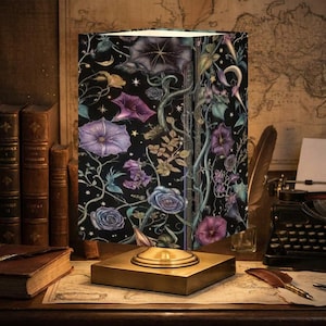 May include: A decorative lamp with a black shade featuring a floral design with purple and blue flowers, vines, and bats. The lamp sits on a gold-colored square base. The background includes books, a map, and a typewriter.