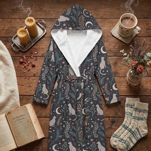 May include: A dark gray hooded robe featuring a repeating pattern of light gray cats, crescent moons, stars, and botanical sprigs. The robe has a white interior lining, long sleeves, and a matching fabric tie belt at the waist for closure.