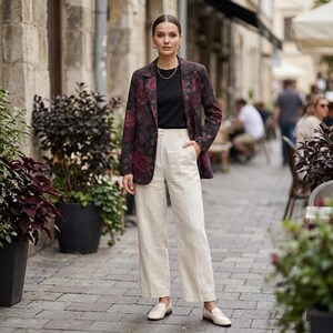 May include: A woman wearing a burgundy blazer with a floral pattern, a black top, and cream-colored wide-leg pants. She is also wearing cream-colored loafers and gold jewelry. The background is a cobblestone street with potted plants.