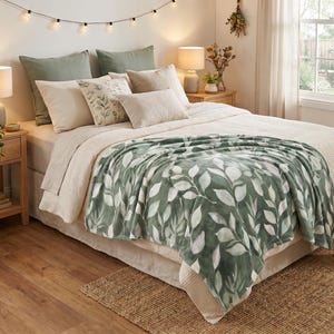 Op de afbeelding: Een zachte plaid met een botanisch bladpatroon in donkergroen en wit, gedrapeerd over een bed. Het bed is opgemaakt met een lichtbeige, gestructureerde sprei, twee grote saliegroene kussens, twee beige kussens en een klein vierkant kussen met bladprint.