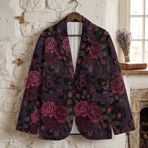 May include: A dark purple blazer with a floral rose pattern in shades of burgundy and pink. The blazer has a classic two-button closure, notched lapels, and two front pockets. The jacket is hanging on a wooden hanger.