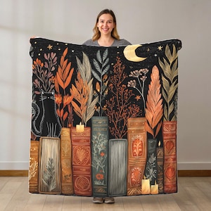 Könnte beinhalten: Eine schwarze Decke mit einer fantastischen Illustration einer schwarzen Katze, Büchern, Pflanzen, einem Mond und Sternen. Das Design verwendet Orange, Braun und Gold vor einem dunklen Hintergrund. Die Decke wird von einer Person gehalten.