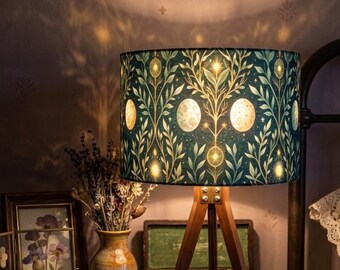 May include: A tripod floor lamp with a cylindrical shade featuring a dark green background, a repeating pattern of light-colored moon phases, and stylized botanical leaf motifs. The lamp emits a warm glow, casting light patterns on the wall.
