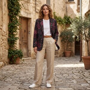 May include: A woman wearing a floral blazer with a white t-shirt, tan pants, and white sneakers. The blazer has a dark background with large red rose patterns. The pants are high-waisted and wide-legged. A brown belt is worn at the waist.