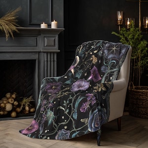 Op de afbeelding: Een zwarte sprei met een bloemenpatroon in paars, blauw en goud, gedrapeerd over een witte fauteuil. Het ontwerp van de sprei bevat wijnranken en sterren tegen een donkere achtergrond. De fauteuil staat in een kamer met een open haard en kaarsen.