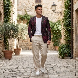 May include: A person wearing a burgundy blazer with a rose pattern, a white t-shirt, and tan pants. The blazer has a dark background with red roses. The pants are a light tan color. The person is wearing white sneakers.