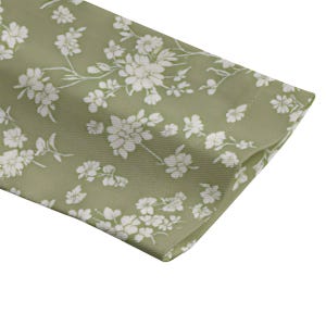 May include: Close-up of a section of fabric in a soft green hue, adorned with a delicate white floral pattern. The fabric is folded, revealing the texture and design of the material. The floral design is a repeating pattern.