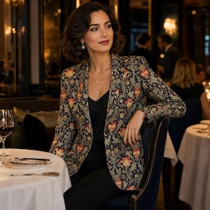 May include: A tailored blazer featuring a dark navy blue base with an intricate floral pattern in shades of orange, yellow, and green. The garment has long sleeves, a notched lapel, and a single-button closure, worn over a simple black top.