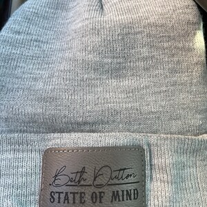 May include: Gray beanie with a brown leather patch that says "Beth Dutton State of Mind" in black lettering.