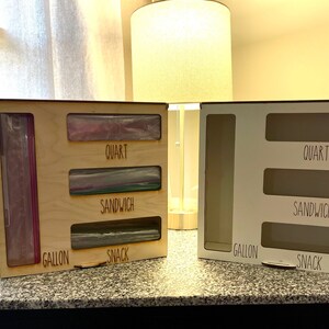 May include: Two wooden storage organizers with labeled compartments for different sizes of plastic bags. The organizer on the left is made of natural wood and has bags in the compartments. The organizer on the right is white and empty. The labels read "Gallon", "Snack", "Sandwich", and "Quart".