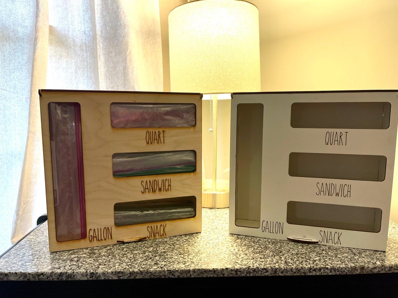 May include: Two wooden storage organizers with labeled compartments for different sizes of plastic bags. The organizer on the left is made of natural wood and has bags in the compartments. The organizer on the right is white and empty. The labels read "Gallon", "Snack", "Sandwich", and "Quart".
