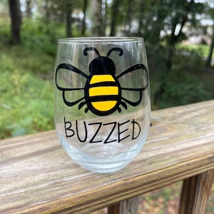 May include: A clear glass wine glass with a black and yellow bee design and the word "buzzed" printed on the bottom.