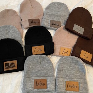 May include: Ten personalized knit beanies with leather patches. The patches feature names like "Hayes", "Jenna", "Austin", "Caleb", "Andy", "Ky", "Kalani", and "Caroline". One beanie has a brown patch with an American flag and the text "Let's Go Brandon".