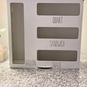 May include: White wooden storage organizer with four compartments labeled "Gallon", "Snack", "Sandwich", and "Quart".