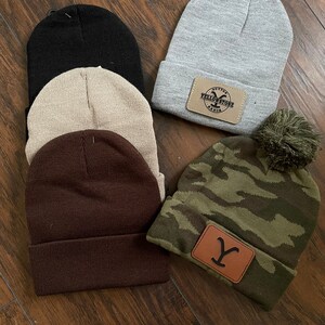 May include: Five different knit beanies with various colors and designs. One beanie is gray with a brown leather patch that says "Dutton Yellowstone Ranch". Another beanie is brown. One beanie is beige. One beanie is black. The last beanie is camouflage green with a brown leather patch with a "Y" on it.
