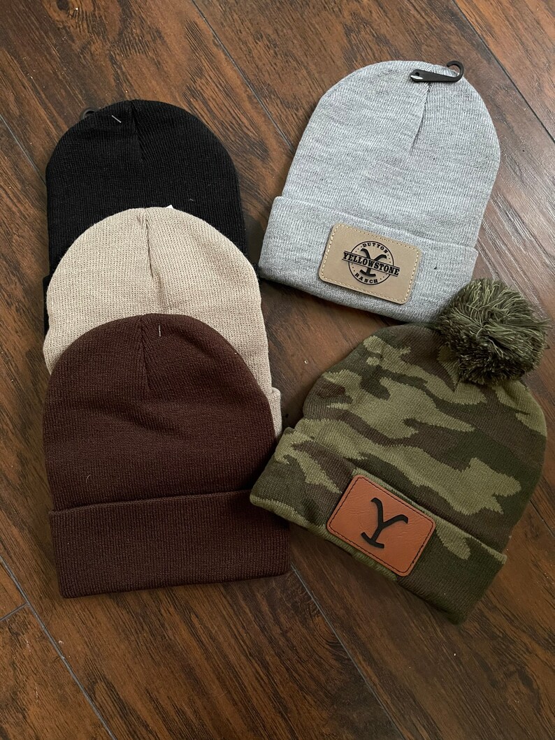 May include: Five different knit beanies with various colors and designs. One beanie is gray with a brown leather patch that says "Dutton Yellowstone Ranch". Another beanie is brown. One beanie is beige. One beanie is black. The last beanie is camouflage green with a brown leather patch with a "Y" on it.