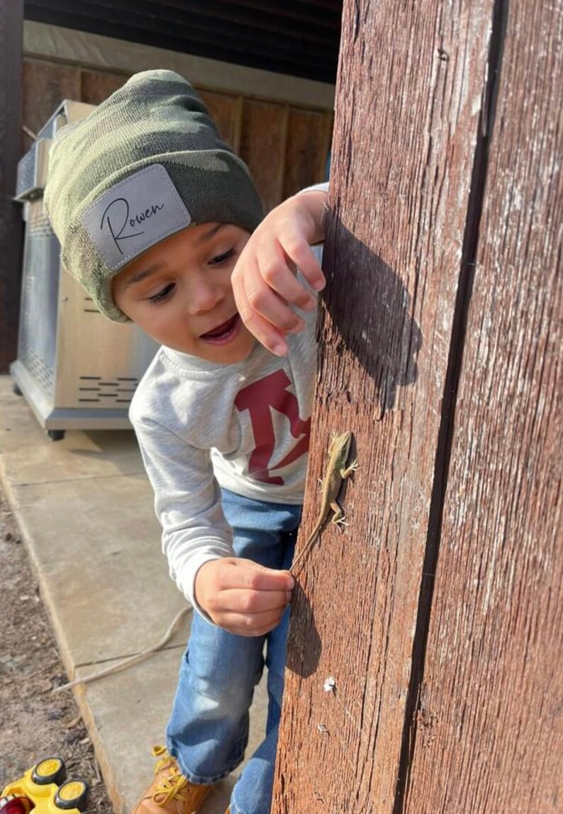 May include: A young boy wearing a green camouflage beanie with the name "Rowan" on it, looks at a small lizard on a wooden surface.