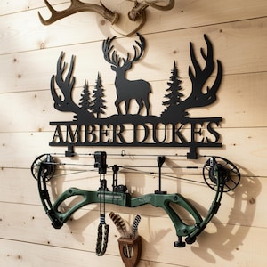 Puede incluir: Decoración de pared de metal negro con una silueta de ciervo, árboles y el nombre "AMBER DUKES", montada en una pared de madera. Debajo, se muestra un arco compuesto verde, con un pequeño soporte de madera con plumas.