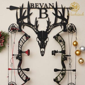 May include: Black metal wall art featuring a deer skull and antlers, with the name "BEVAN" above. Two compound bows are displayed below, one black and red, the other green and black. Decorative lights and ornaments are also present.