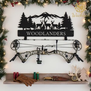 May include: A black metal sign with the word "WOODLANDERS" and a silhouette of a mountain scene with deer and trees. Below the sign is a camouflage compound bow. A shelf displays hunting accessories, including gloves, shells, and a knife.