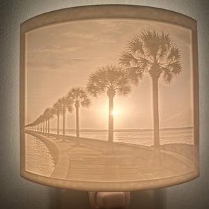 May include: A night light with a beige lampshade featuring a scenic image of a boardwalk lined with palm trees at sunset. The light is plugged into a wall outlet.
