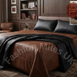 May include: A bed featuring a brown leather frame with a grid-stitched pattern, two black leather pillows, and a black leather throw blanket. A circular logo in the corner displays the text Fashion Miles UK and Genuine Leather.