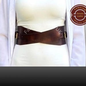 May include: A wide dark brown leather waist belt featuring a crisscross design that wraps around the midsection. The belt includes two metallic buckles on the sides and is worn over a white top. A circular logo reads Fashion Miles UK Genuine Leather.
