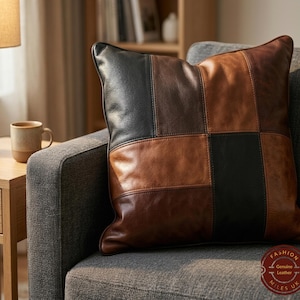 Op de afbeelding: Een vierkant leren kussen met een patchwork ontwerp in bruin- en zwarttinten ligt op een grijze bank. Het kussen heeft een getextureerd uiterlijk en is waarschijnlijk gevuld met een zacht materiaal voor comfort en ondersteuning.