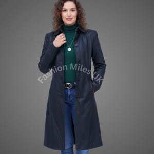 May include: A long, navy blue leather coat with a button-up front and a collared neckline. The coat is worn over a green turtleneck and blue jeans. A silver necklace and a black belt with a gold buckle complete the look. The coat appears to be knee-length.