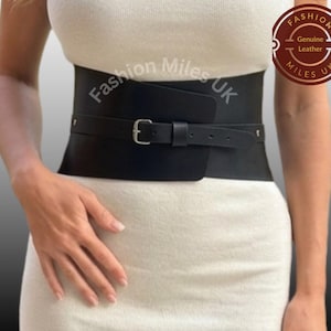 May include: A wide black leather corset belt featuring a silver-toned metal buckle and a thin strap overlay. The belt is worn over a white sleeveless top. The text Fashion Miles UK is visible on the belt and in a circular logo on the top right.
